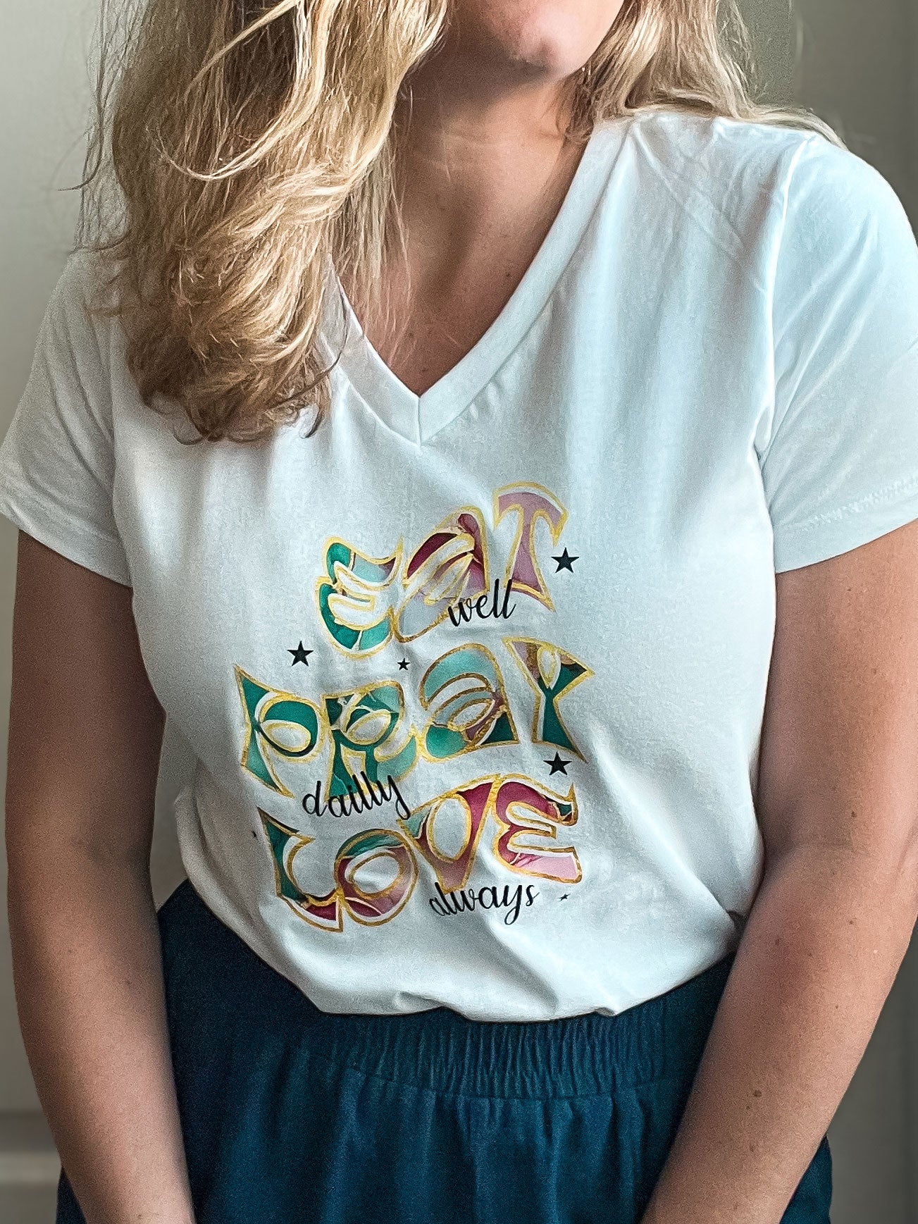 Woman wearing a white V-neck T-shirt with the phrase “Eat well, Pray daily, Love always” in bold, colorful letters with a fun, whimsical style. The words are outlined in gold with playful patterns inside each letter, paired with small black stars.