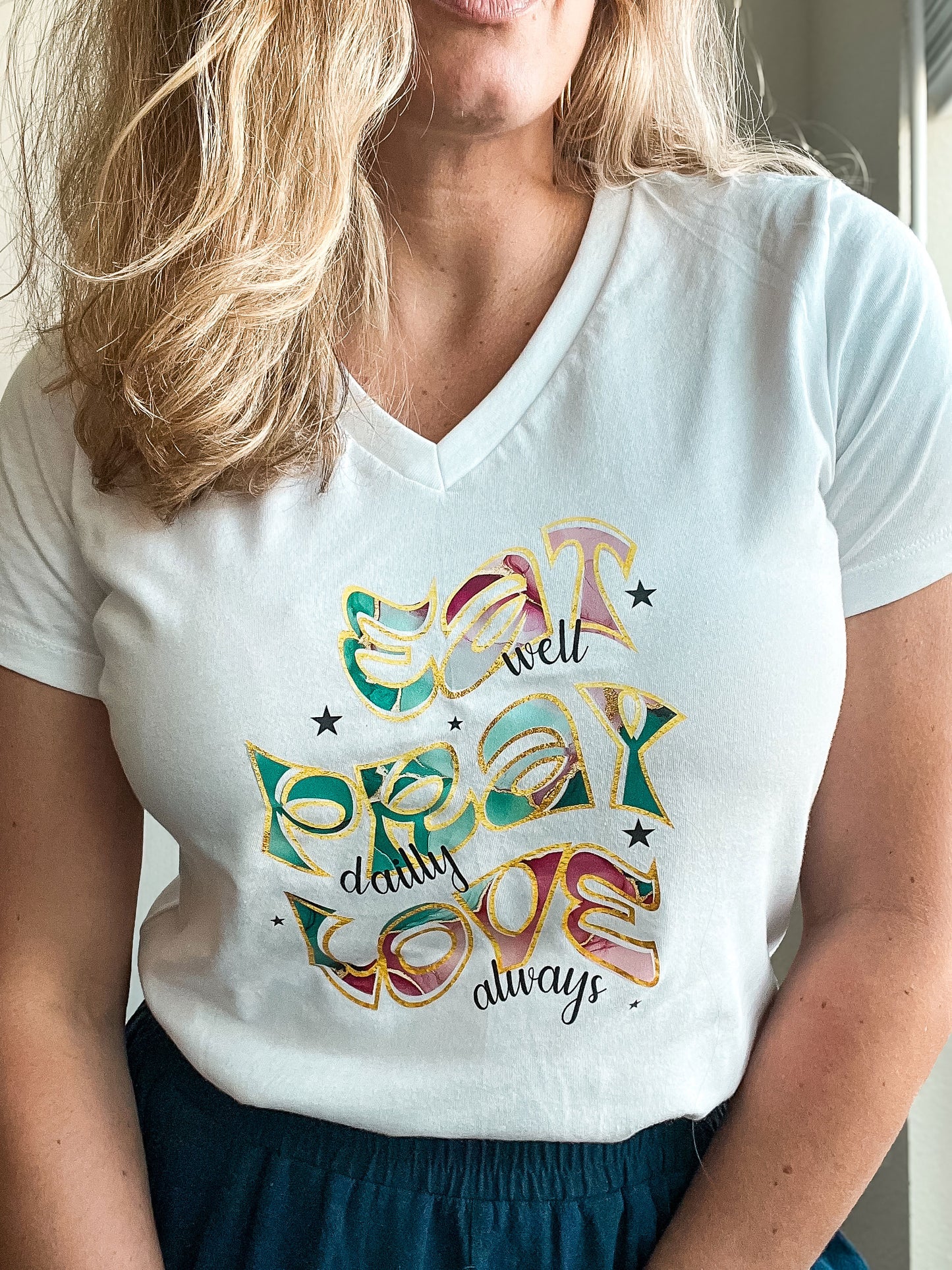 Close-up of a white V-neck T-shirt featuring the text “Eat well, Pray daily, Love always” in large, multicolored and gold-outlined letters. The artistic font and scattered stars create a joyful and faith-inspired message.