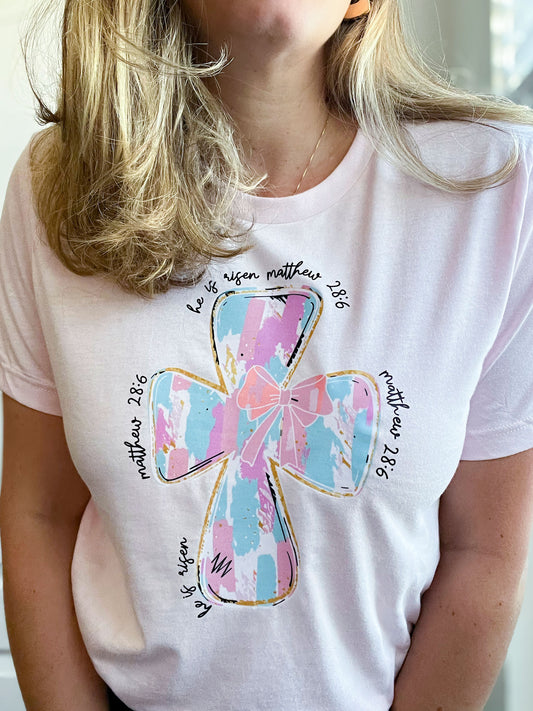Woman wearing a light pink T-shirt featuring a large pastel cross design in shades of blue, pink, and white with gold outlines. Surrounding the cross, the text reads ‘He is risen’ and ‘Matthew 28:6’ in a handwritten-style font.