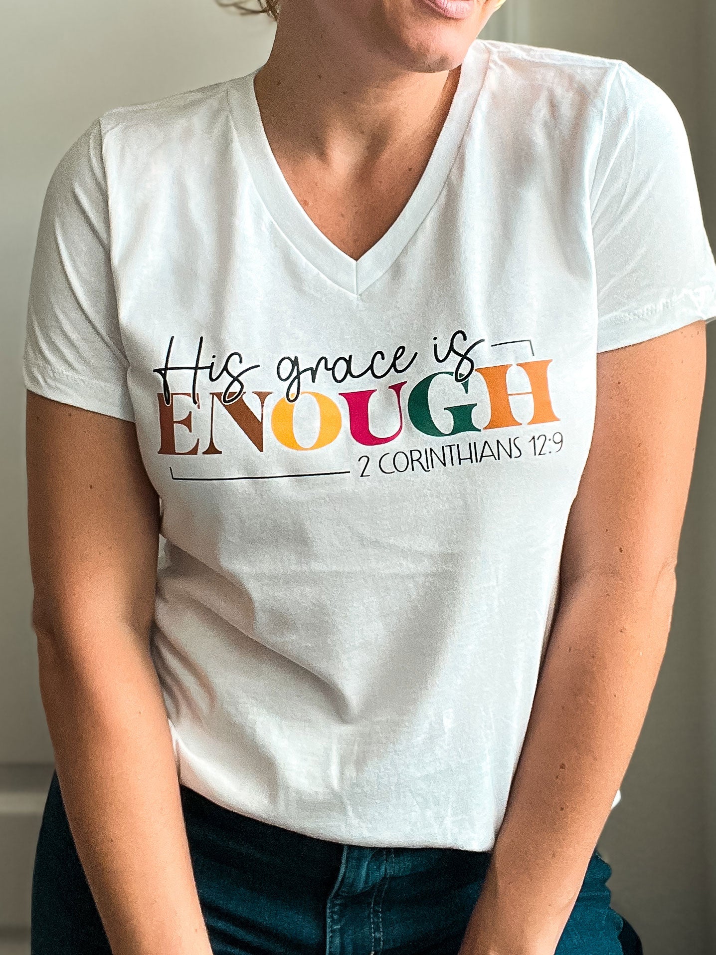 Woman wearing a white V-neck T-shirt with the phrase “His grace is ENOUGH – 2 Corinthians 12:9” printed in black and colorful lettering. The word “ENOUGH” stands out in bold rainbow colors, creating an uplifting and faith-filled look.