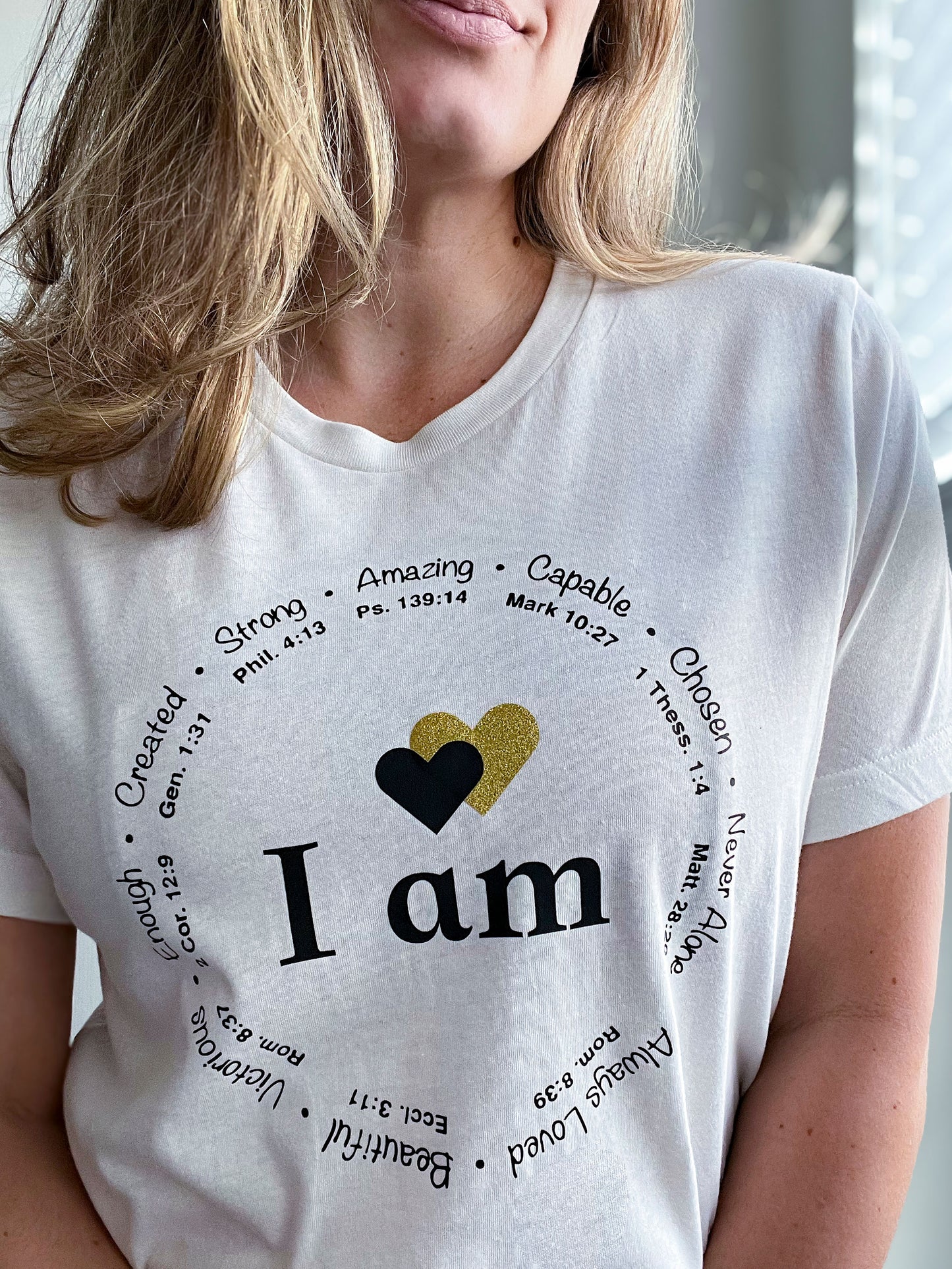 Close-up of a woman wearing a vintage white T-shirt featuring the phrase “I am” in bold black letters, with two hearts above—one black and one gold glitter. Surrounding the text is a circular arrangement of affirmations like “Strong,” “Capable,” “Created,” “Victorious,” and “Always Loved,” each paired with Bible verses. The design reflects faith-based encouragement and empowerment, part of Spark of Faith’s handmade apparel collection.
