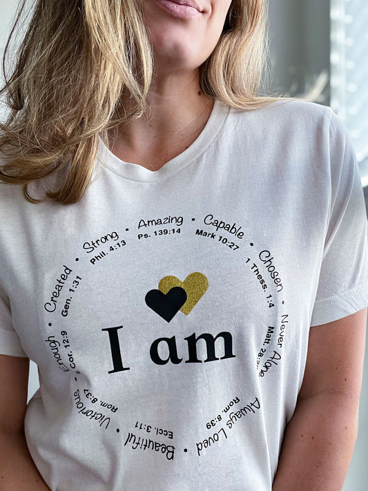 Close-up of a woman wearing a vintage white T-shirt featuring the phrase “I am” in bold black letters, with two hearts above—one black and one gold glitter. Surrounding the text is a circular arrangement of affirmations like “Strong,” “Capable,” “Created,” “Victorious,” and “Always Loved,” each paired with Bible verses. The design reflects faith-based encouragement and empowerment, part of Spark of Faith’s handmade apparel collection.