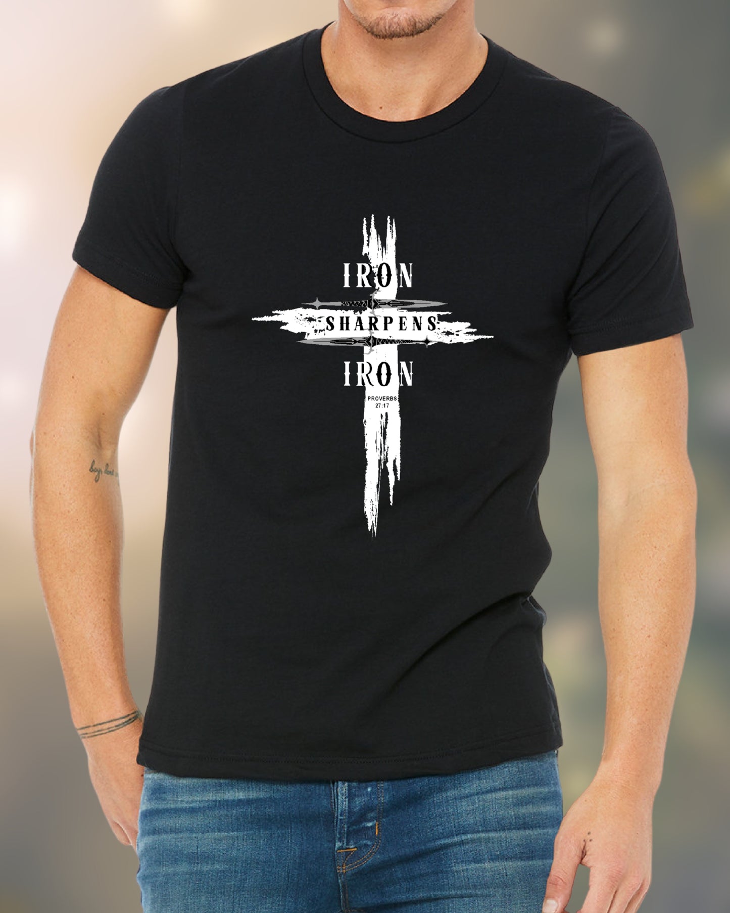 Man wearing a black T-shirt featuring a bold distressed cross design with the words ‘IRON SHARPENS IRON’ in white capital letters. Below the text, ‘Proverbs 27:17’ is printed. Styled with blue jeans.