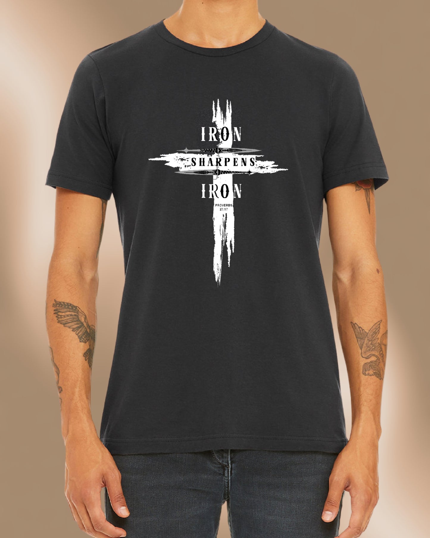Man wearing a dark grey T-shirt featuring a bold distressed cross design with the words ‘IRON SHARPENS IRON’ in white capital letters. Below the text, ‘Proverbs 27:17’ is printed. Styled with black jeans.
