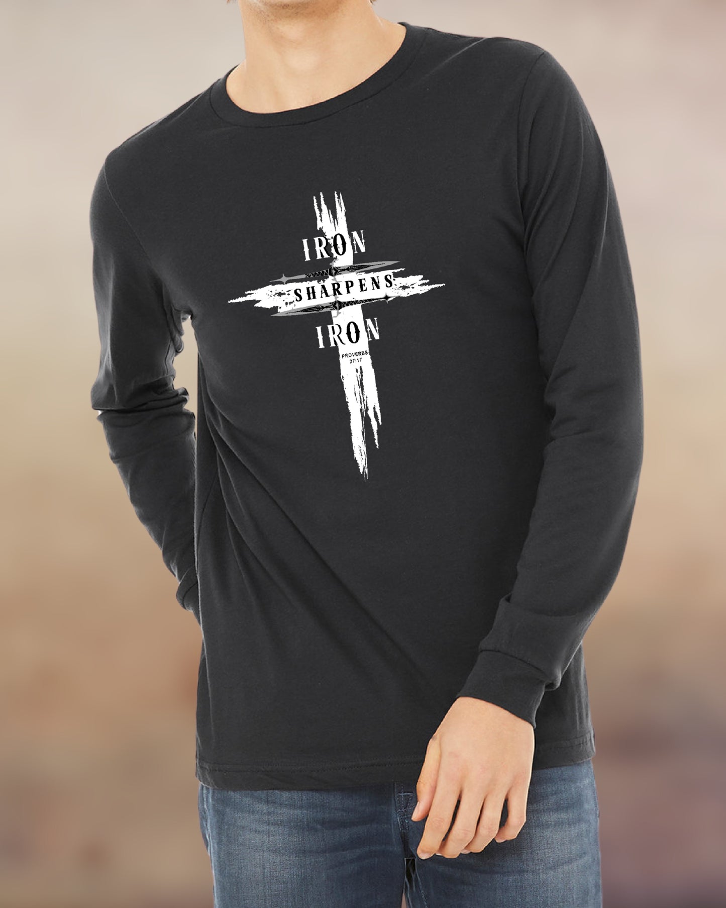 Man wearing a dark grey long-sleeve shirt with a bold white cross design that reads “Iron Sharpens Iron” from Proverbs 27:17. The graphic features a distressed brushstroke effect, giving it a rugged and powerful look suitable for faith-filled men who lead and uplift others.