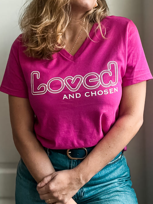 Woman wearing a bright pink V-neck T-shirt with ‘Loved and Chosen’ printed in white. The word ‘Loved’ features a playful outline. Styled with high-waisted blue jeans, a black belt with a gold buckle, and a delicate gold necklace.