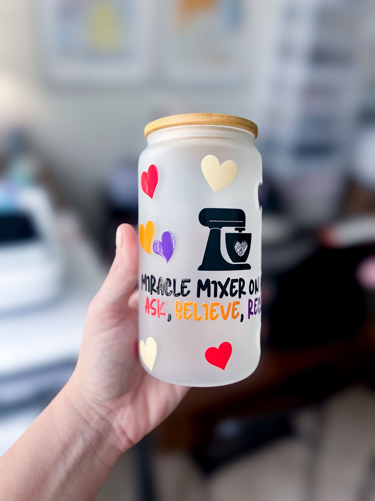 Hand holding a frosted glass cup with a bamboo lid, decorated with colorful heart designs and a black stand mixer illustration. The text reads ‘MIRACLE MIXER ON’ followed by ‘ASK, BELIEVE, RECEIVE’ in a mix of black, red, orange, and purple lettering. Blurred background of a workspace with framed artwork and crafting supplies.