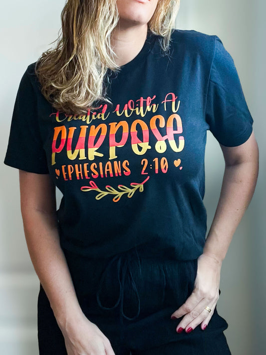 Woman wearing a black T-shirt that reads “Created with a Purpose – Ephesians 2:10” in colorful gradient text. The design is accented with small hearts and a leafy branch, giving a warm, bold message of faith and identity.