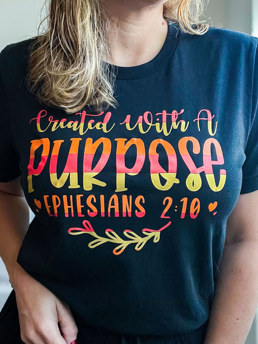 Close-up of a woman wearing a black T-shirt with the bold phrase “Created with a Purpose – Ephesians 2:10” in red, orange, and yellow gradient lettering. Decorative hearts and a leafy branch underline the verse, making this a vibrant faith statement.