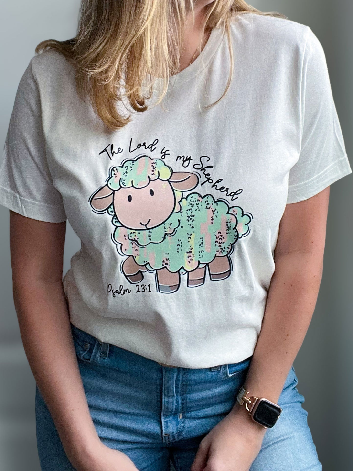 Woman wearing a white T-shirt featuring a cute pastel-colored cartoon sheep with the text “The Lord is my Shepherd” above it and “Psalm 23:1” below. The sheep is illustrated in soft pink, mint green, and gold brushstroke textures, creating a playful and faith-filled design.