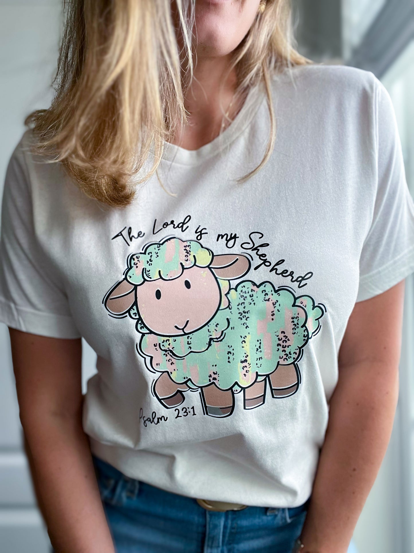 Close-up of a white T-shirt with a charming cartoon sheep in pastel hues and the verse “The Lord is my Shepherd – Psalm 23:1.” The colorful, textured design adds a fun and comforting feel to this faith-based tee.
