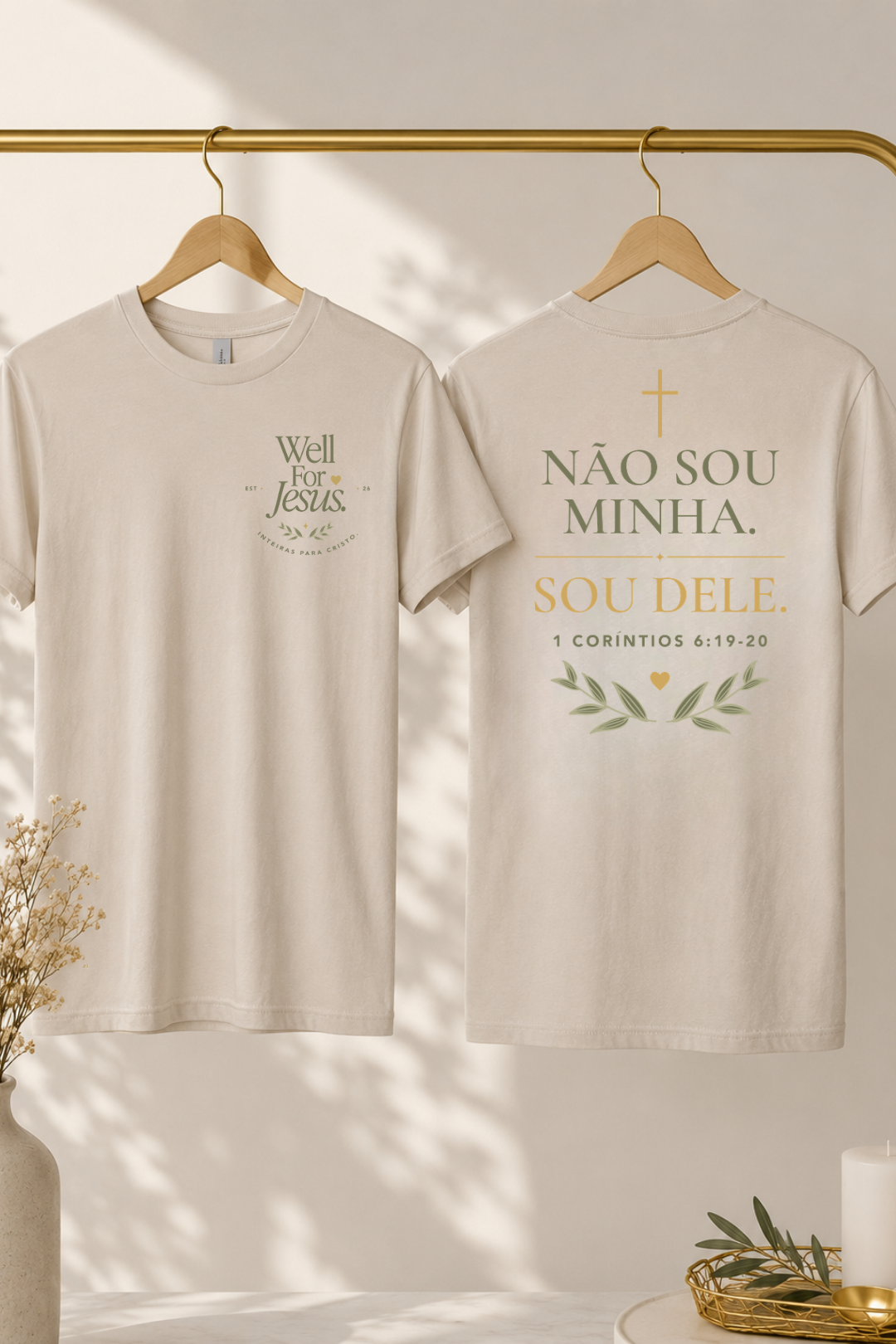 Two cream-colored t-shirts hanging on wooden hangers from a gold clothing rack. The front of the left shirt features a green graphic reading 'Well For Jesus', and the back of the right shirt features a gold and green graphic reading 'NÃO SOU MINHA. SOU DELE"
