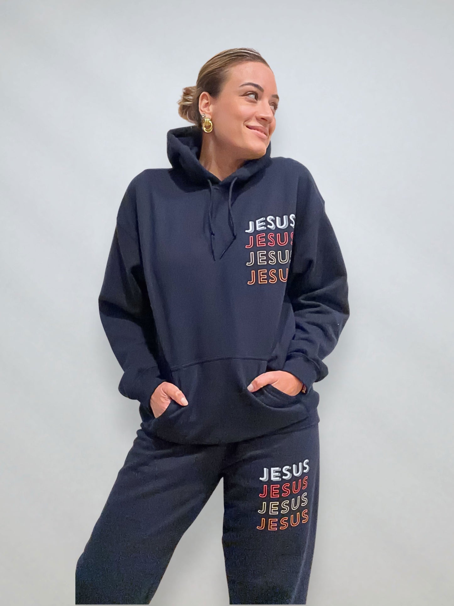 Woman wearing navy JESUS Repeat hoodie and sweatpants set, hands in pockets, front design on chest and leg.