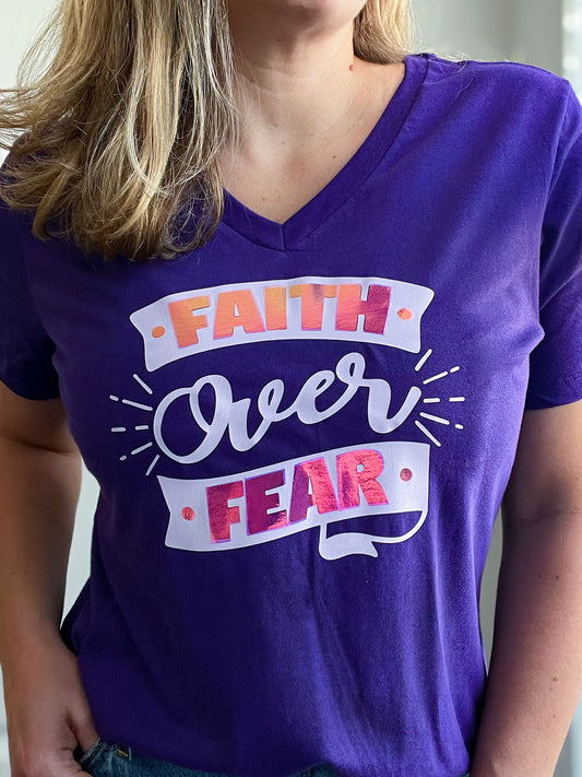 Close-up of a woman wearing a deep purple V-neck T-shirt with the phrase “Faith Over Fear” in bold, decorative lettering. The design features light purple script with holographic pink elements, giving it a reflective, eye-catching effect. The faith-inspired shirt is part of Spark of Faith’s handmade apparel collection.
