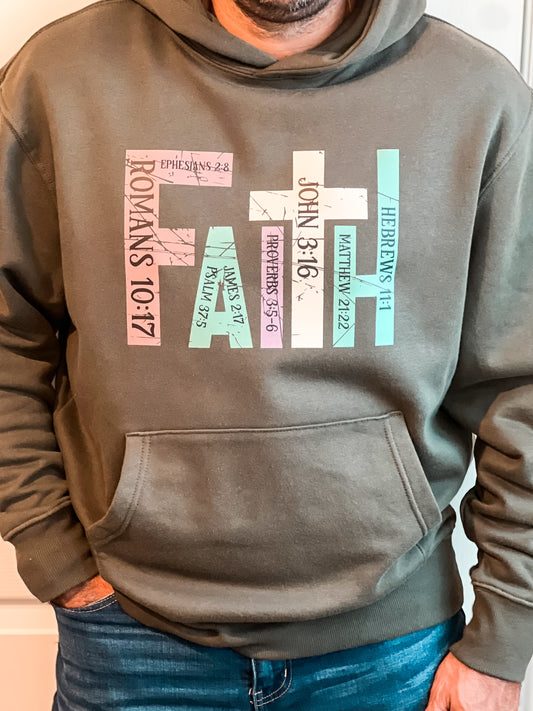 Man wearing a military green hoodie with the word “FAITH” printed in block letters across the chest. Each letter is filled with a Bible verse reference, including Romans 10:17, Ephesians 2:8, Psalm 37:5, Proverbs 3:5–6, John 3:16, Matthew 21:22, and Hebrews 11:1. The design combines bold typography and scripture for a powerful, faith-centered statement.