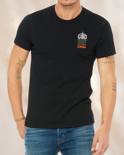 Front view of a black t-shirt with colorful brushstroke-style design reading “God Has a Plan – Trust It, Live It, Enjoy It” and a cross inside the letter “O” in “God.”