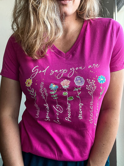  Woman wearing a bright pink V-neck T-shirt with floral illustrations and the inspirational phrase “God says you are” followed by positive affirmations next to each flower: “Unique, Special, Lovely, Precious, Strong, Chosen, Forgiven.” The shirt features colorful flower designs aligned with each word.