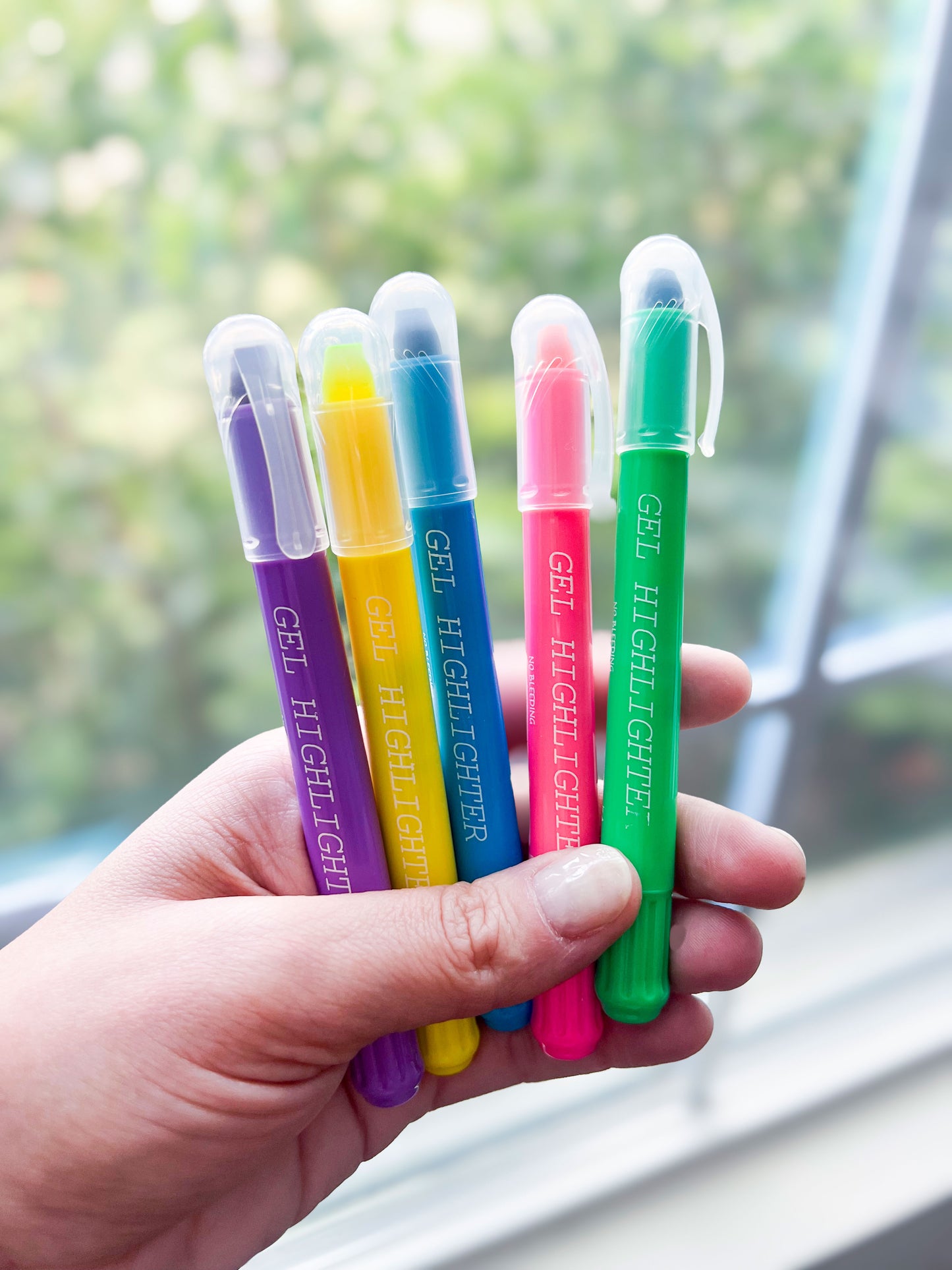 Hand holding a set of five colorful gel highlighters in purple, yellow, blue, pink, and green. Each highlighter has a clear cap and ‘GEL HIGHLIGHTER’ printed on the body. The background features a softly blurred window view with greenery.