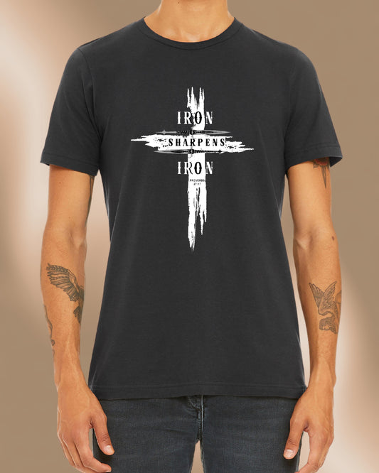 Man wearing a dark grey T-shirt featuring a bold distressed cross design with the words ‘IRON SHARPENS IRON’ in white capital letters. Below the text, ‘Proverbs 27:17’ is printed. Styled with black jeans.
