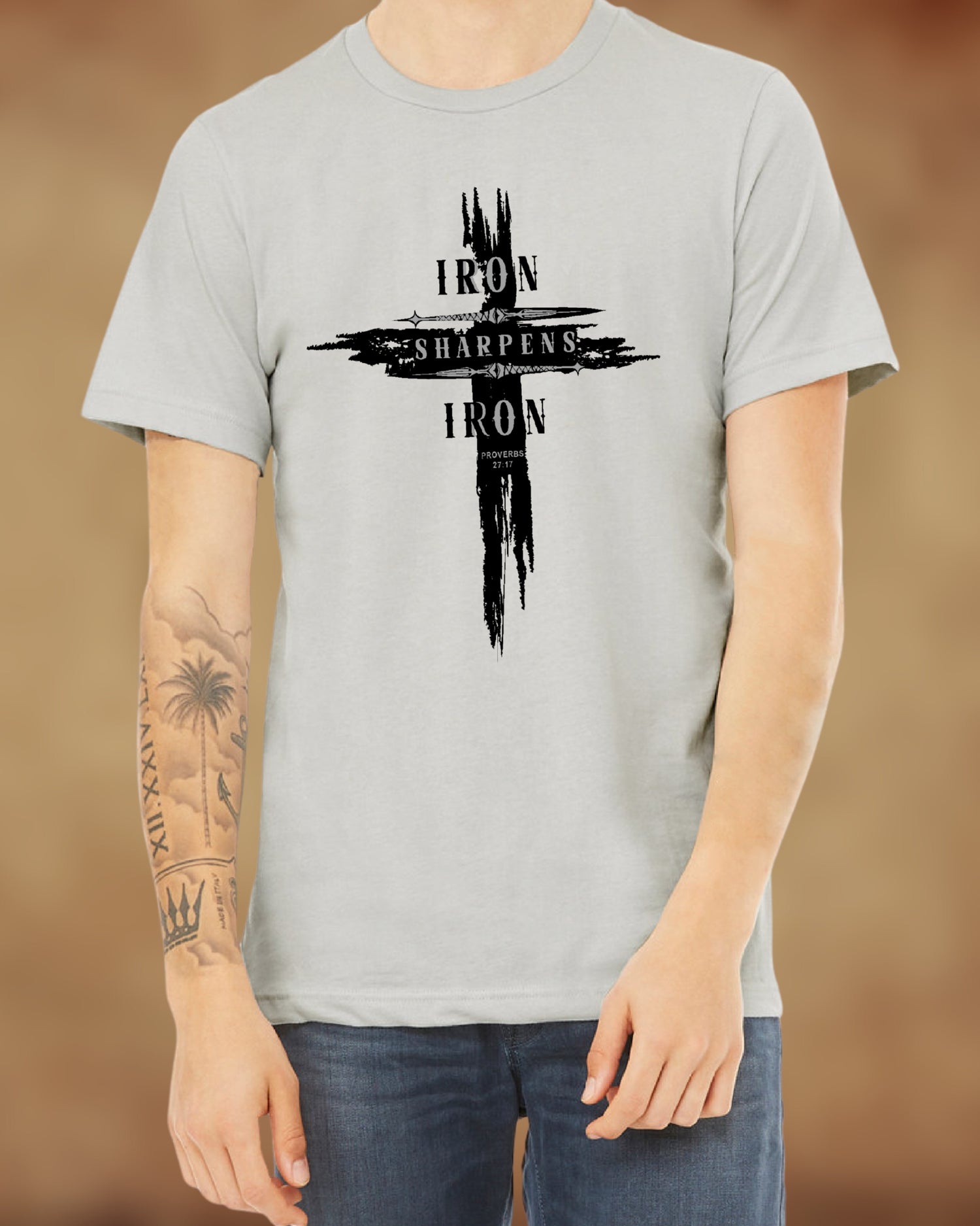 Man wearing a light gray T-shirt featuring a distressed cross design with the words ‘IRON SHARPENS IRON’ in bold black capital letters. Below the text, ‘Proverbs 27:17’ is printed. Styled with blue jeans.