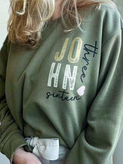 Front of a military green sweatshirt with a creative layout of the words “JOHN three sixteen” in distressed block and cursive fonts, with a small pink heart accent. A soft and stylish expression of one of the Bible’s most beloved verses.