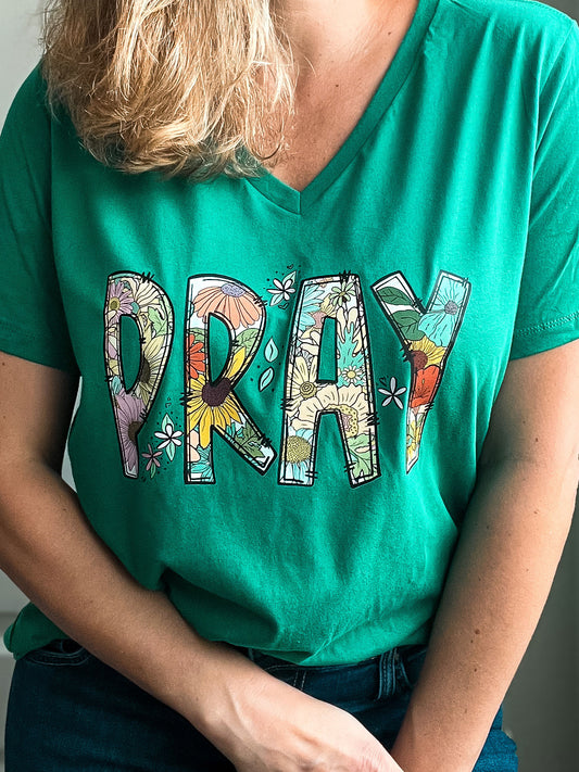 Woman wearing a green V-neck T-shirt with the word “PRAY” in large, bold letters filled with colorful floral patterns. The shirt combines faith and cheerful design, creating a bright and uplifting message.