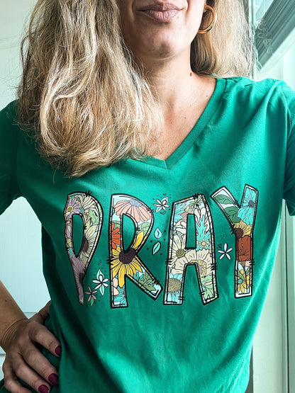 Close-up of a woman wearing a green V-neck T-shirt with the word “PRAY” in large floral-patterned letters. The vibrant design features a mix of colorful flowers inside each letter, conveying a joyful and faith-filled message.