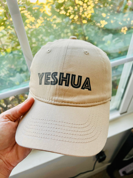 Close-up of a beige baseball cap with the word “YESHUA” printed in bold black letters across the front. The hat is held up by a hand near a sunlit window with greenery in the background, emphasizing its casual and faith-centered design.