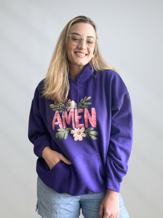 Person wearing a purple hoodie with 'AMEN' and floral designs on a plain background