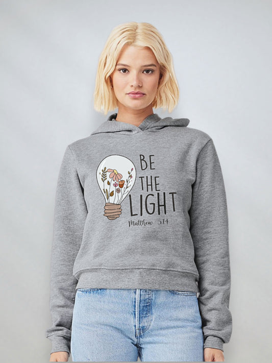 Woman wearing an athletic heather Christian hoodie with a light bulb design and the phrase “Be the Light” from Matthew 5:14
