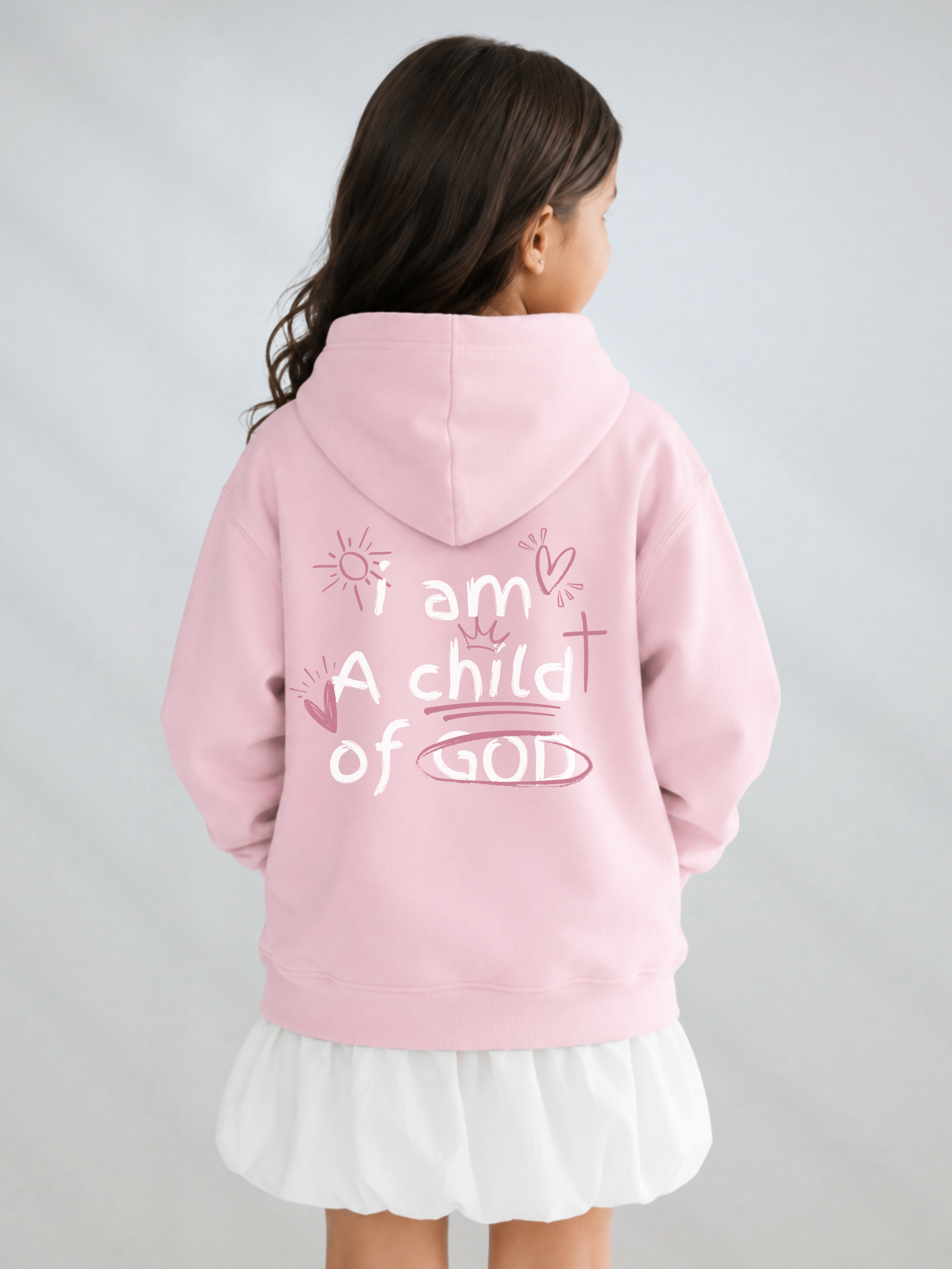 A young girl viewed from behind wearing a light pink hoodie with the text 'i am A child of GOD' printed in white on the back, paired with a white ruffled skirt, against a light gray studio background.