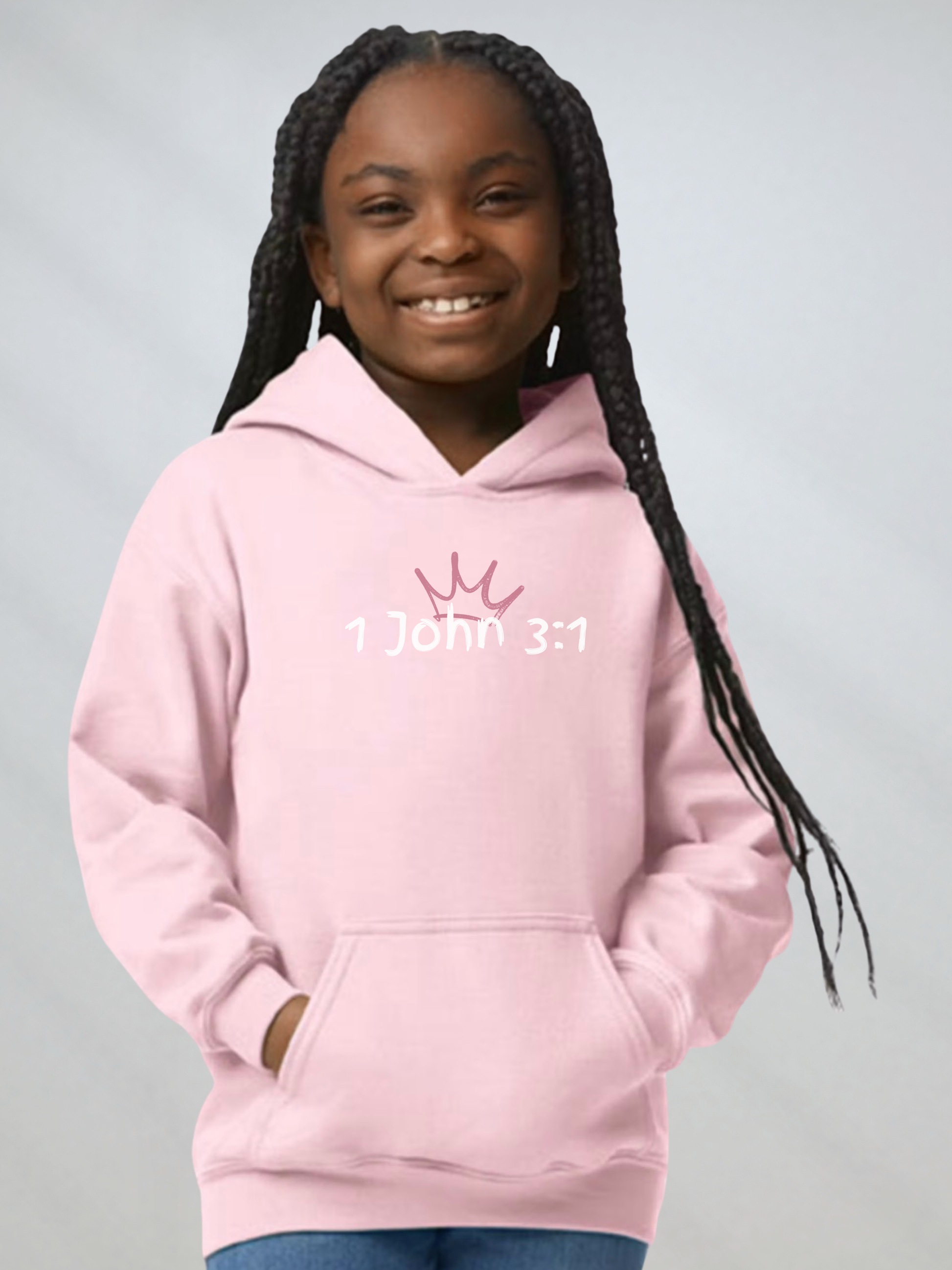 A young girl with long black braids smiling while wearing a light pink hoodie with a white crown graphic and the text '1 John 3:1' printed on the chest, set against a plain light gray background.