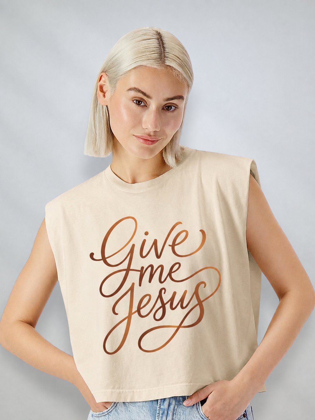 A blonde woman wearing a beige sleeveless crop top with 'Give Me Jesus' printed in brown cursive script, paired with light blue jeans, against a light gray studio background.