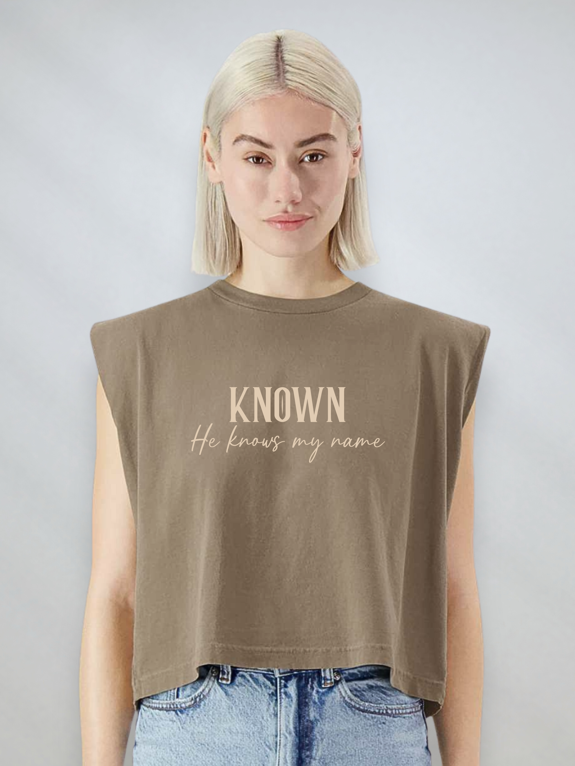 A blonde woman wearing a cropped sleeveless t-shirt in faded brown with the text 'KNOWN He knows my name' printed on the front, paired with light wash blue jeans, against a light gray studio background.