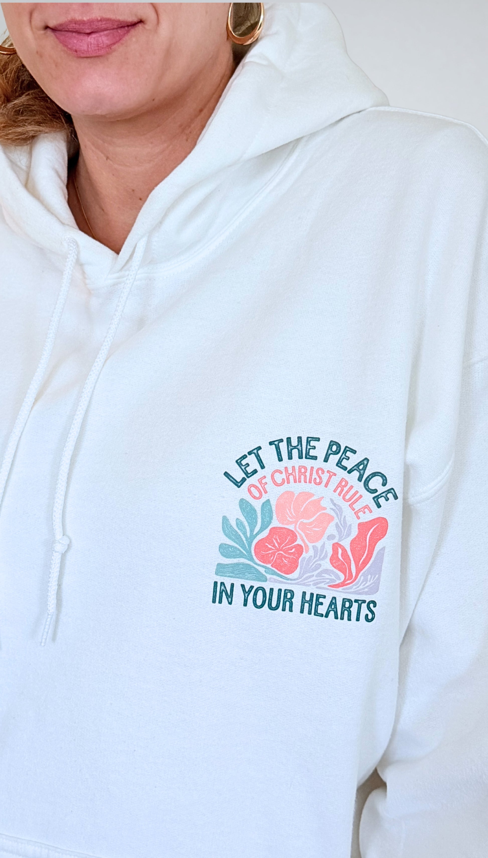 Close-up of white hoodie featuring floral design and Christian verse "Let the Peace of Christ Rule in Your Hearts."