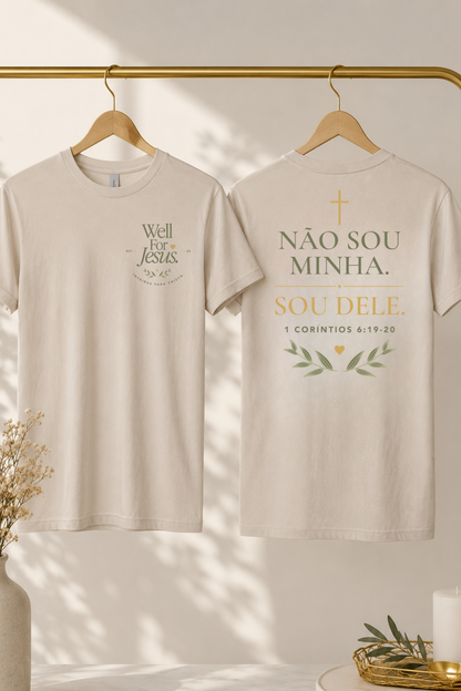 Two cream-colored t-shirts hanging on wooden hangers from a gold clothing rack. The front of the left shirt features a green graphic reading 'Well For Jesus', and the back of the right shirt features a gold and green graphic reading 'NÃO SOU MINHA. SOU DELE"